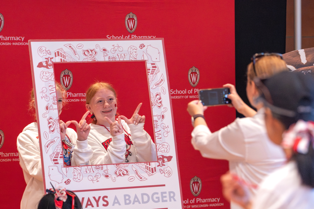 A red-haired girl in a white sweatshirt poses for a photo inside a large, rectangular photo frame prop that reads "ALWAYS A BADGER." She is making the 'W' hand sign. A person's hand is visible on the right, taking her photo with a smartphone. The background is the red School of Pharmacy backdrop.