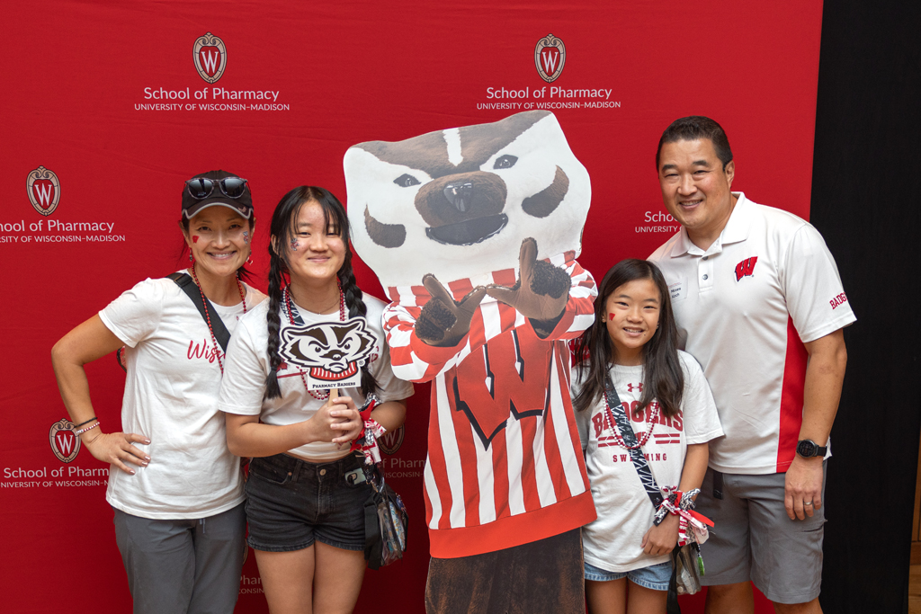A family of four, including two adults and two children wearing red and white University of Wisconsin-Madison apparel, poses with a large cardboard cutout of Bucky Badger. The backdrop is red, featuring the School of Pharmacy University of Wisconsin-Madison logo.