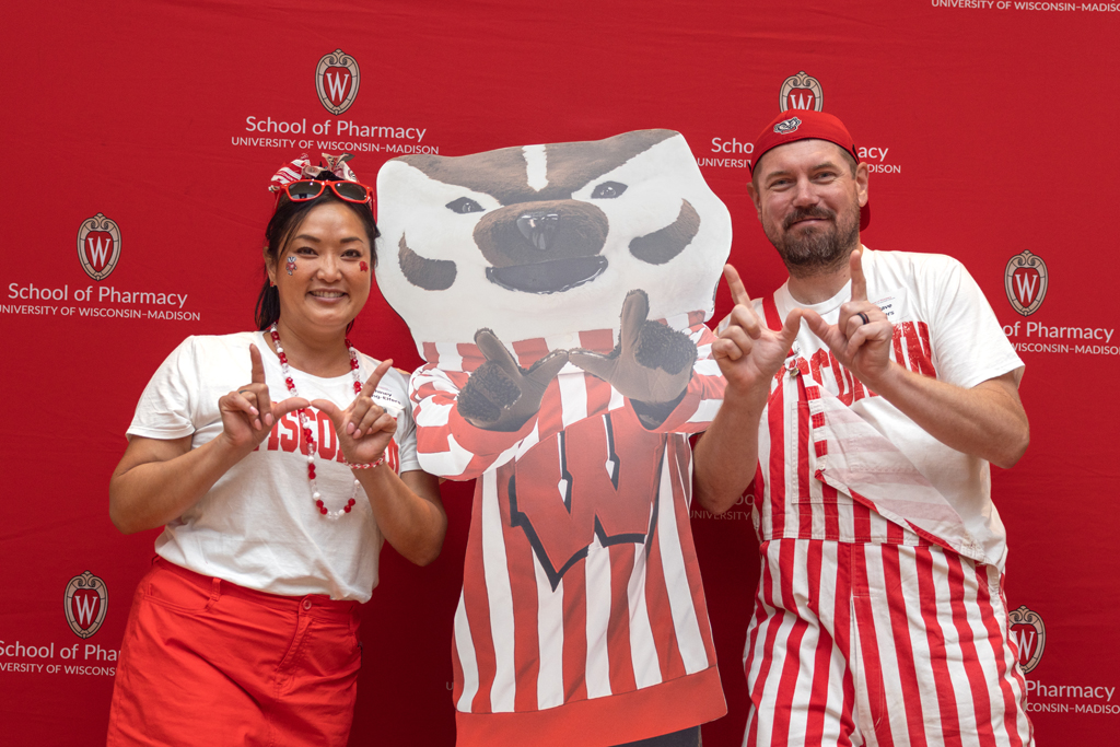 A woman in a red skirt and white shirt and a man in red and white striped overalls pose with a cardboard cutout of Bucky Badger. They are standing in front of the red School of Pharmacy backdrop and both are making the 'W' hand sign.