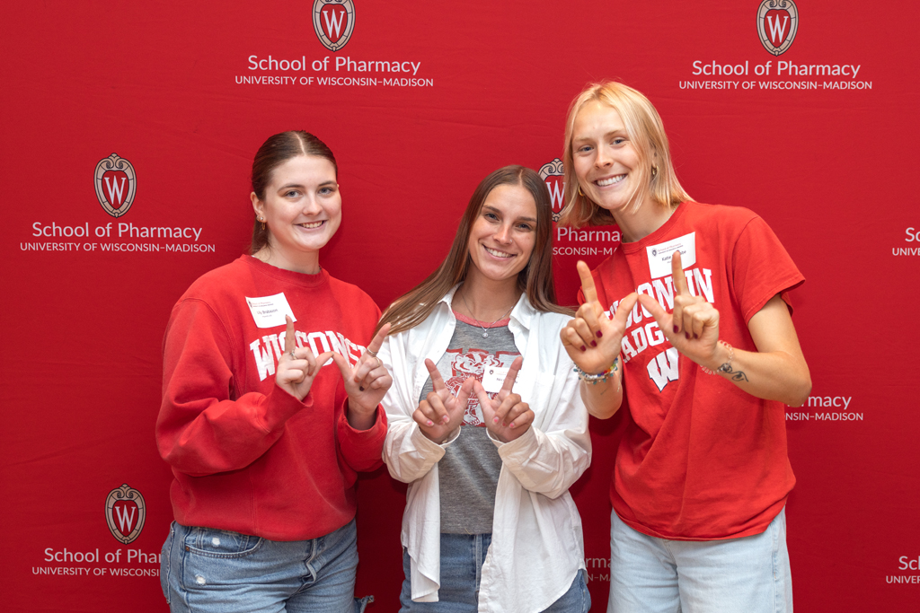 Three young women, two wearing red Wisconsin shirts and one wearing a gray shirt and white button-down, stand together in front of the red University of Wisconsin-Madison School of Pharmacy backdrop, all making the 'W' hand sign.