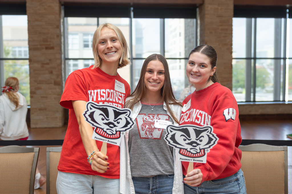 phar092025-069.jpg: Three young women stand together, smiling in an indoor event space with large windows. The women on the left and right are wearing red shirts and holding small black and white signs shaped like the Bucky Badger logo that say "PHARMACY BADGERS."