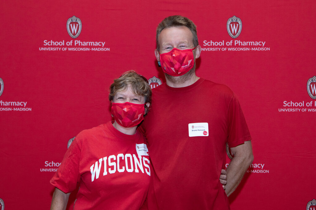 Two pharmacy alumni standing in front of red School of Pharmacy backdrop