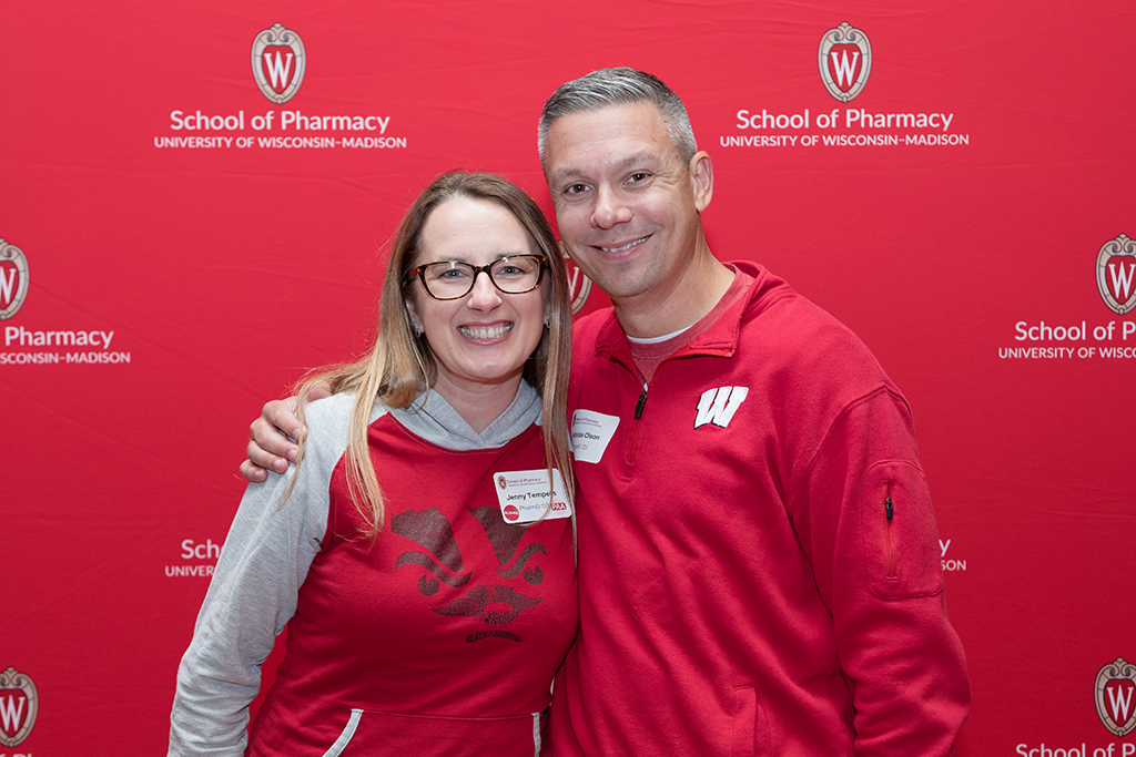 Alumni smiling in front of red School of Pharmacy backdrop