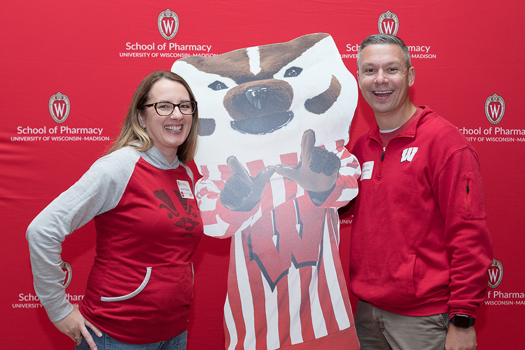 Alumni posing next to Bucky Badger cardboard cutout in front of red School of Pharmacy backdrop