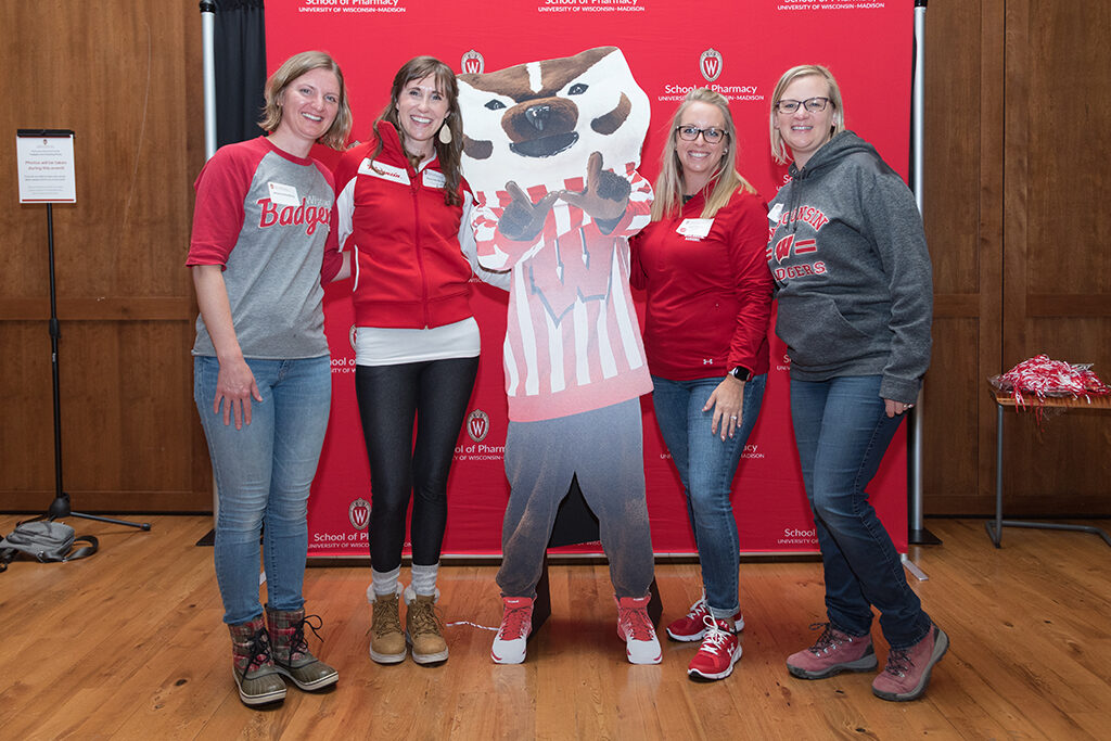Alumni pose with Bucky cardboard cutout in front of red School of Pharmacy logo background