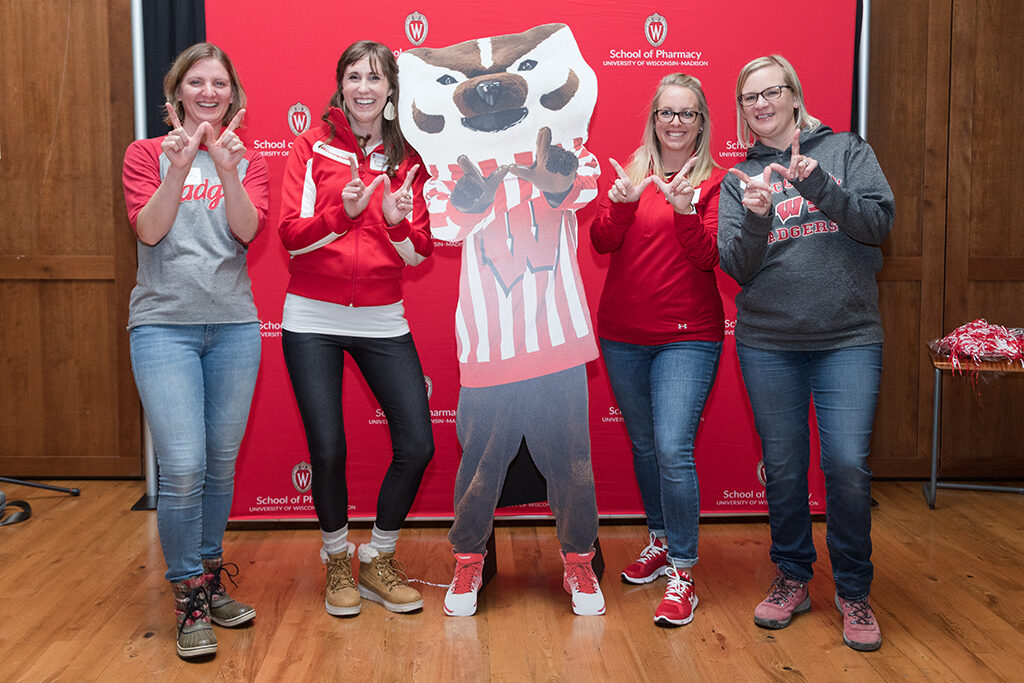 Alumni hold up "W"s with Bucky cardboard cutout in front of red School of Pharmacy logo background