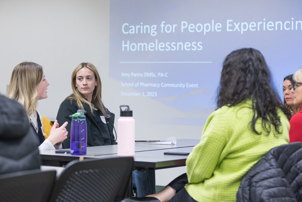 PharmD students talking around a table, with a Caring for People Experiencing Homelessness presentation on the wall behind them.